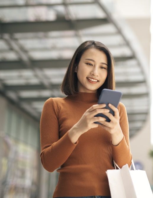 Portrait of elegant young Asian woman holding shopping bags and using smartphone on the go while leaving mall
